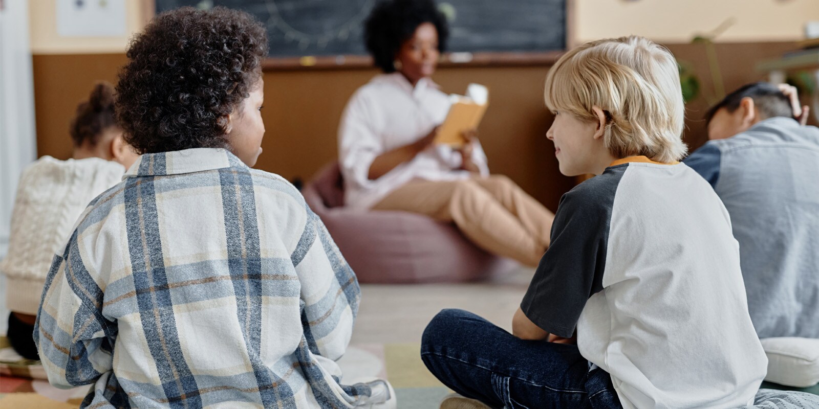 Students smile sitting in classroom while listening to a teacher read in the background