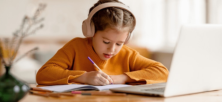 Child wearing headphones, using a laptop and writing on a notepad.